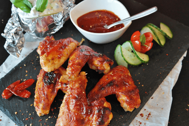 Glazed chicken wings with red sauce on a slate platter, served with cucumber, cherry tomatoes, dip, and extra sauce.