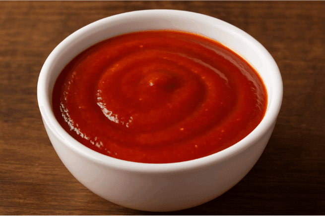 A small white bowl filled with a thick, smooth red sauce, swirled neatly at the surface, placed on a wooden table.