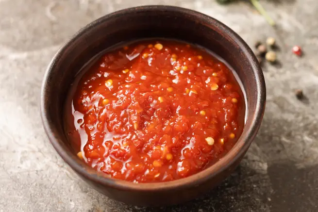 Bowl of chunky red salsa with visible chili seeds, served in a rustic brown bowl on a stone surface.