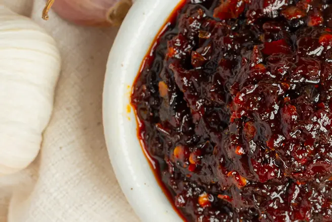 Close-up of homemade crunchy shallot and garlic chili oil in a ceramic bowl, highlighting its rich, spicy texture with visible chili flakes and garlic pieces.
