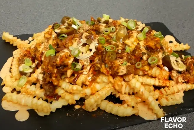 "Plate of crinkle-cut Papas locas fries topped with creamy sauce, spicy chili, sliced jalapeños, and chopped green onions served on a black slate board.